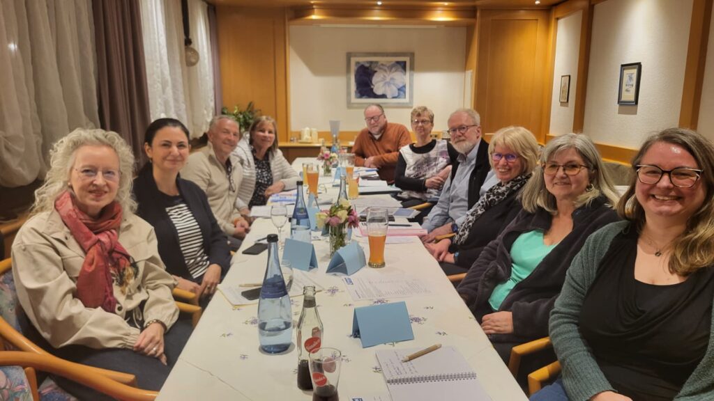Eine Gruppe von zwölf Erwachsenen, sechs Männer und sechs Frauen, sitzen in einem warm beleuchteten Raum um einen langen Tisch, schauen in die Kamera und lächeln. Der Tisch ist mit Getränken, Zeitungen, Blumen und Notizblöcken gedeckt.