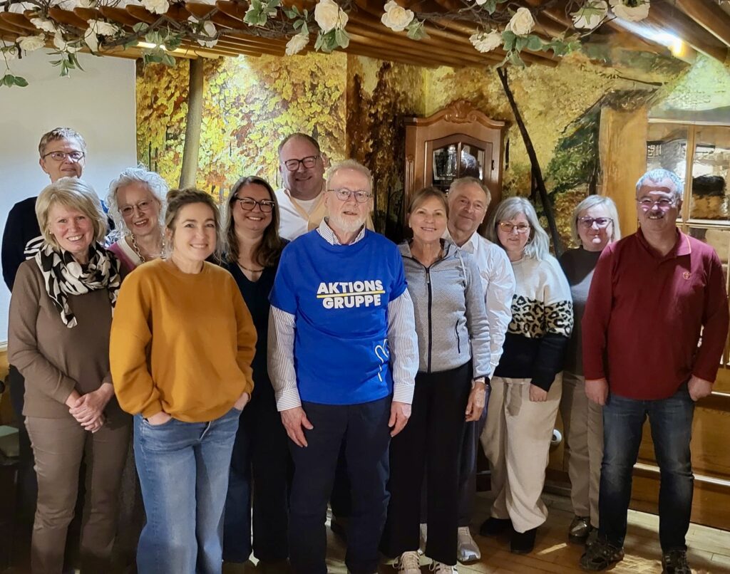 Eine Gruppe von zwölf Erwachsenen, die lächelnd in einem gemütlichen, warm beleuchteten Raum mit Holzbalken und Wandmalereien zum Thema Natur stehen. Eine Person in der Mitte trägt ein blaues Hemd mit der Aufschrift AKTIONS GRUPPE.