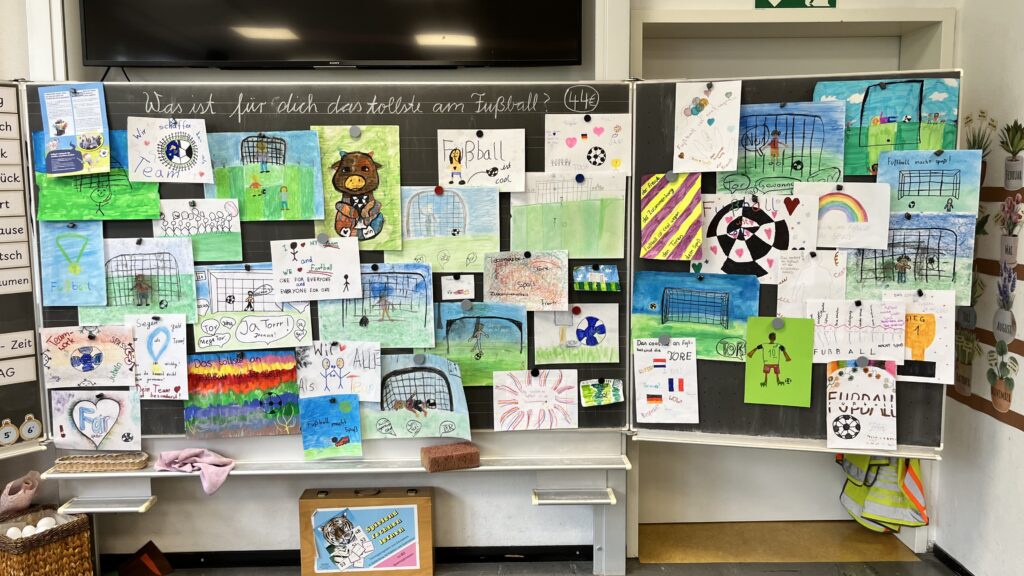 Eine Kreidetafel im Klassenzimmer ist mit bunten Kinderzeichnungen bedeckt, die sich hauptsächlich um Fußball, Tore, Bälle und jubelnde Fans drehen. Auf der Tafel oben wird auf Deutsch gefragt, was die Schülerinnen und Schüler am meisten am Fußball mögen. Darunter befinden sich Kunst- und Schulsachen.